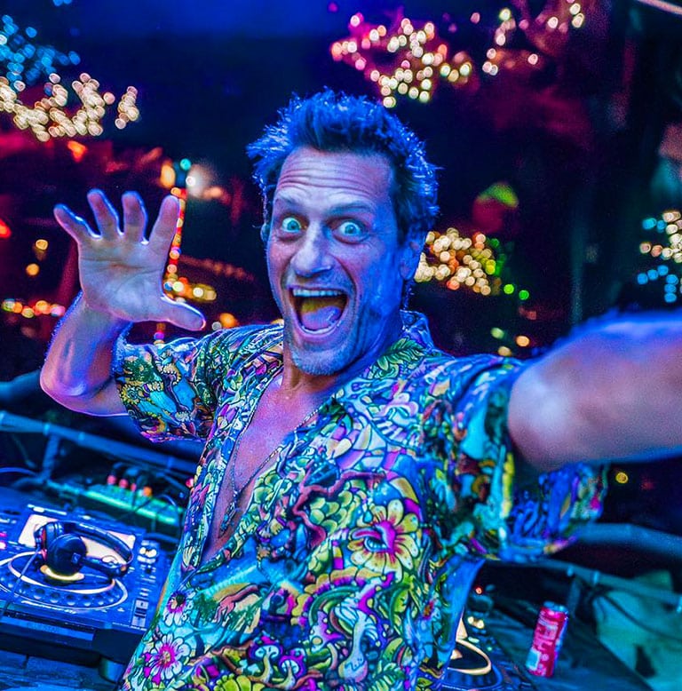DJ mit buntem Hemd, der an einem Mischpult steht und in die Kamera lächelt.