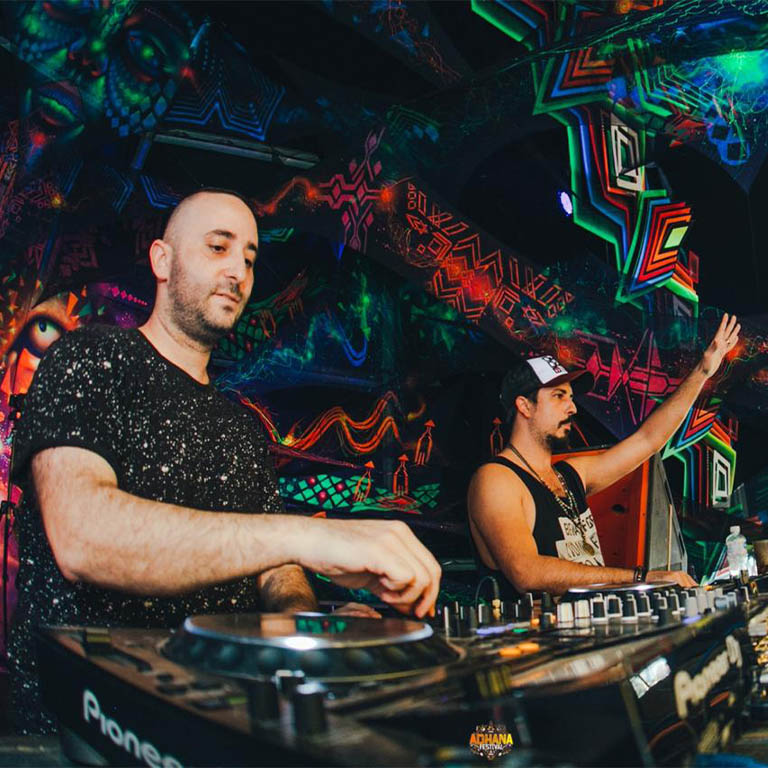 Zwei DJs stehen an einem Mischpult mit buntem, psychedelischem Hintergrund während einer Live-Performance.