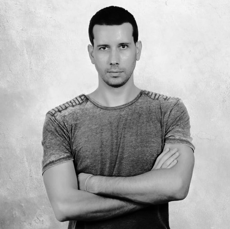 Porträt eines Mannes mit verschränkten Armen, der in einem grauen T-Shirt vor einem neutralen Hintergrund steht.