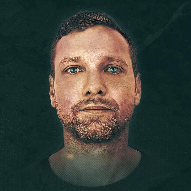 Porträt eines Mannes mit blauen Augen und Bart, vor einem dunklen Hintergrund. Der Gesichtsausdruck ist neutral.