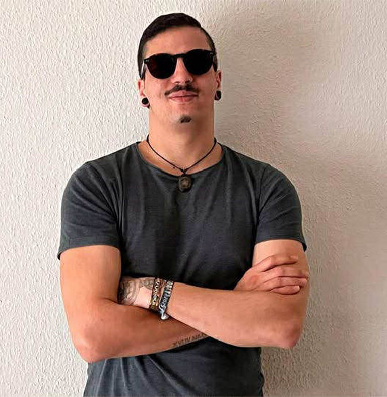 Mann mit Sonnenbrille, T-Shirt und Armbändern, der mit verschränkten Armen posiert.