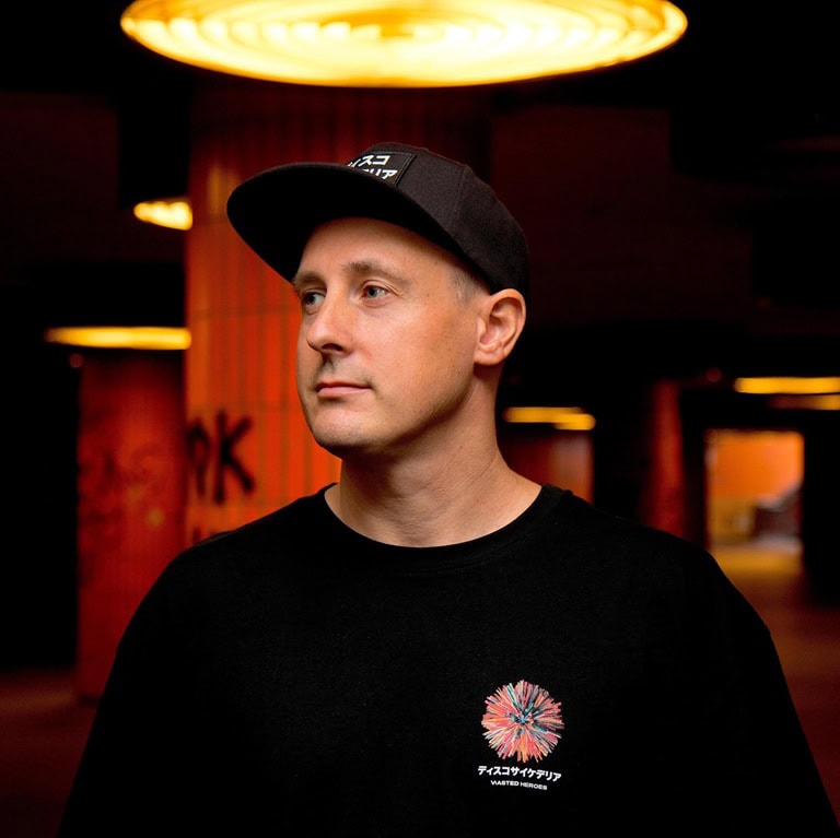 Mann mit schwarzem T-Shirt und Cap, der in einem urbanen Raum steht, mit beleuchteten Säulen im Hintergrund.